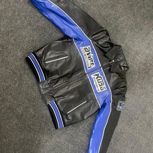 以下が含まれることがあります： 白のアクセントが付いた黒と青のレーシングジャケット。 ジャケットはジッパーフロント、スタンドカラー、袖と胸に青いストライプが特徴です。 ジャケットには「Avire」と「Xuss」の文字がプリントされています。