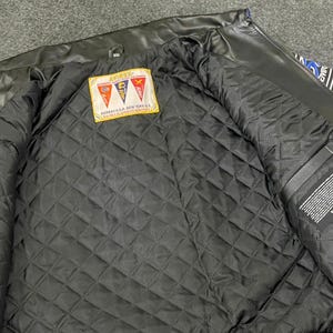 以下が含まれることがあります： 黒い革のジャケット、キルティング加工された黒い裏地付き。 ジャケットの内側にはテキスト入りのラベルと、赤、黄、青の旗のパッチが付いています。 ジャケットは開いており、内部の詳細が明らかになっています。