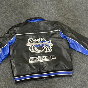 以下が含まれることがあります： 黒い革のジャケット。袖と襟に青色のアクセント。背中には「Black Knights」と「AVIREX」の文字が入った白と青の刺繍グラフィック。裾は青と白のストライプのリブ編みです。