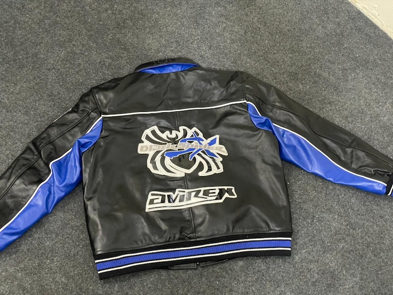 以下が含まれることがあります： 黒い革のジャケット。袖と襟に青色のアクセント。背中には「Black Knights」と「AVIREX」の文字が入った白と青の刺繍グラフィック。裾は青と白のストライプのリブ編みです。