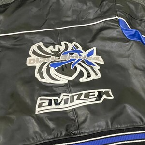 以下が含まれることがあります： 黒い革のジャケットで、青と白のアクセントが付いています。背面には「Black Spider」と「Avirex」の文字が白とシルバーで大きく描かれています。袖には青色のパネルがあります。