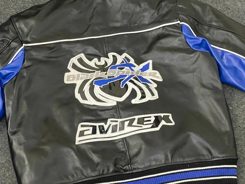 以下が含まれることがあります： 黒い革のジャケットで、青と白のアクセントが付いています。背面には「Black Spider」と「Avirex」の文字が白とシルバーで大きく描かれています。袖には青色のパネルがあります。
