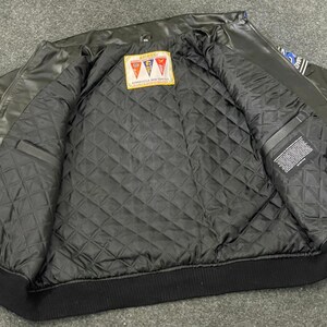 以下が含まれることがあります： 黒い革のジャケットで、青と白のアクセントが付いています。ジャケットはキルティング加工された黒い裏地と黒いリブ編みの裾が特徴です。赤、白、オレンジの形をしたパッチが内側にあります。ジャケットは開いています。