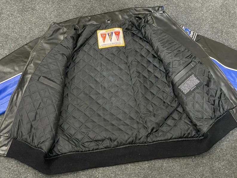 以下が含まれることがあります： 黒い革のジャケットで、青と白のアクセントが付いています。ジャケットはキルティング加工された黒い裏地と黒いリブ編みの裾が特徴です。赤、白、オレンジの形をしたパッチが内側にあります。ジャケットは開いています。