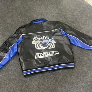 以下が含まれることがあります： 黒い革のジャケットで、袖と襟に青いアクセントがあります。背面には白と青のグラフィックと「Avirex」の文字があります。ジャケットは、青と白のストライプが入ったリブ編みの裾と袖口が付いています。