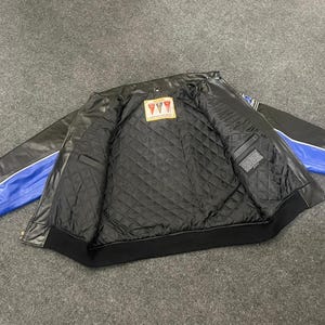 以下が含まれることがあります： 黒い革のジャケットで、袖に青と白のアクセントがあります。内側はキルティング加工された黒い裏地と小さなラベルが付いています。ジャケットは、リブ編みの襟と袖口を備えたクラシックなボンバースタイルです。