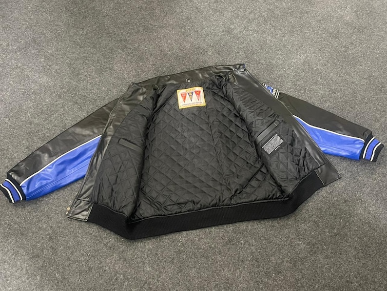 以下が含まれることがあります： 黒い革のジャケットで、袖に青と白のアクセントがあります。内側はキルティング加工された黒い裏地と小さなラベルが付いています。ジャケットは、リブ編みの襟と袖口を備えたクラシックなボンバースタイルです。