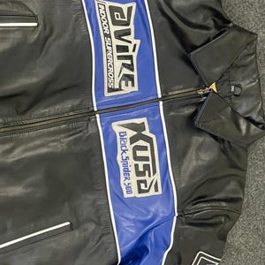 以下が含まれることがあります： 黒い革のジャケットで、青と白のアクセントが施されています。「Avire Indoor Supercross」と「Xusa Black Spider 500」の文字が胸にあります。ジッパー開閉と袖に小さなパッチが付いています。
