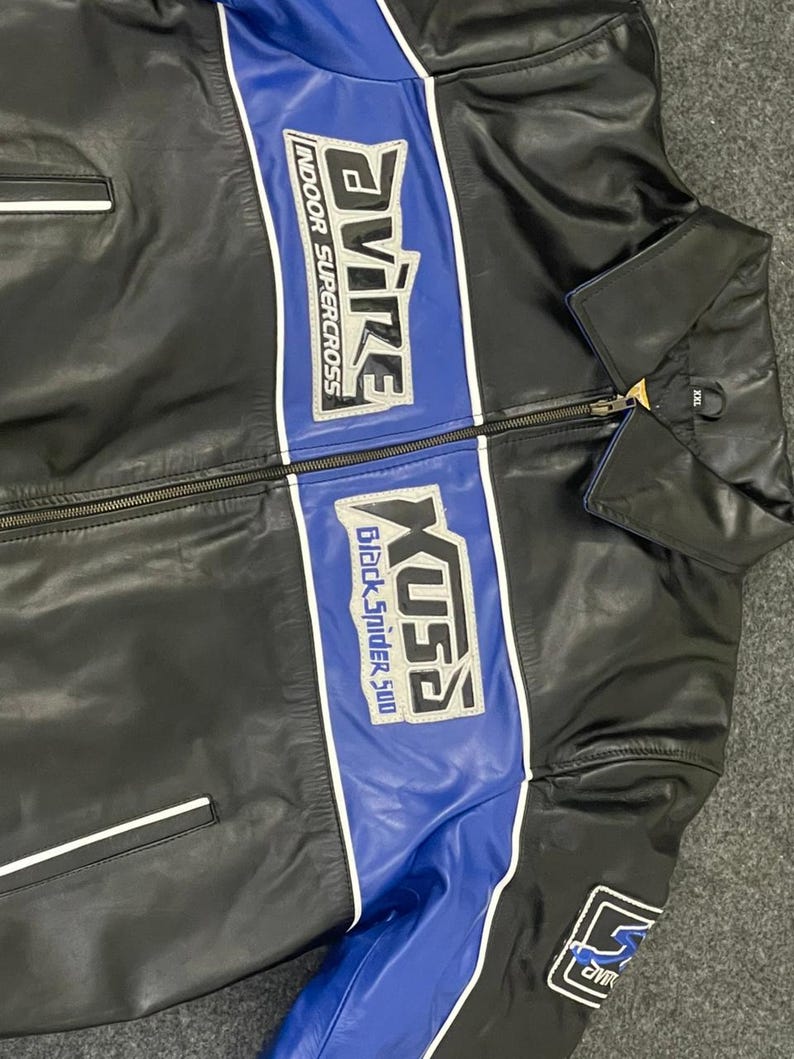 以下が含まれることがあります： 黒い革のジャケットで、青と白のアクセントが施されています。「Avire Indoor Supercross」と「Xusa Black Spider 500」の文字が胸にあります。ジッパー開閉と袖に小さなパッチが付いています。