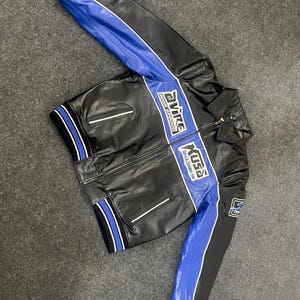 以下が含まれることがあります： 白のアクセントが付いた黒と青のレーシングジャケット。袖と胸には青いストライプがあり、白で「Avire」と「Xuss」の文字が書かれています。ジッパー開閉とリブ編みの袖口が付いています。