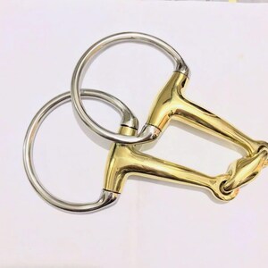 May include: A horse bit with a gold-colored mouthpiece and silver-colored rings. The bit is designed with a jointed mouthpiece and circular rings for attachment to the bridle.