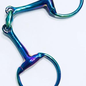 Op de afbeelding: Een paardenbit met een iriserende afwerking. De bit heeft twee D-ringen die verbonden zijn door een centrale schakel. Het metaal reflecteert tinten blauw, groen en paars. Deze paardensportuitrusting is ontworpen voor gebruik met een hoofdstel.