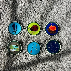 May include: Six circular badges featuring colorful illustrations: a purple lavender sprig on blue, a red ladybug on green, a campfire on dark blue, a treasure map on brown, a fishing rod on blue, and a yellow constellation on a dark blue background.