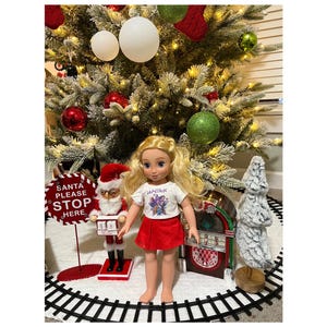 May include: A doll with blonde hair, wearing a white t-shirt and red skirt, is positioned in front of a decorated Christmas tree. A Santa Claus figure, a jukebox, and a sign reading "Santa Please Stop Here" are also visible.