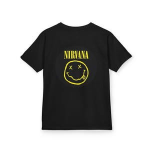 May include: Black t-shirt featuring a yellow Nirvana smiley face logo. The logo includes the band's name above the smiley face. The smiley face has crossed-out eyes and a tongue sticking out.