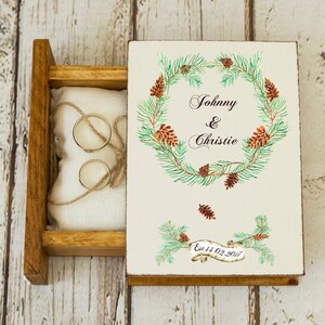 May include: Wooden ring box with a white lid featuring a watercolour wreath of pine branches and pine cones. The lid also has the text "Johnny & Christie Est 14.02.2017". The box is open and a white pillow with two gold rings is inside.