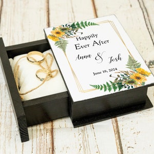 May include: A black wooden ring box with a white lid featuring a floral design with sunflowers and ferns. The lid has a gold frame with the text "Happily Ever After Anna & Josh June 19, 2024".