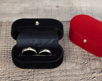 May include: Two gold wedding bands with small blue stones are displayed in a black velvet ring box. A red velvet ring box is also shown.