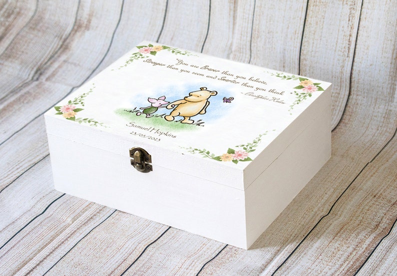 May include: White wooden keepsake box with a hinged lid and metal clasp. The lid features a Winnie the Pooh illustration with Piglet, a butterfly, and a quote. Floral and leaf accents frame the top. The box is personalized with a name and date.