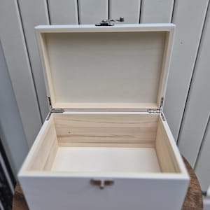 May include: An open, white wooden box with a silver clasp and hinges. The interior of the box is natural wood, contrasting with the painted exterior. The box is sitting on a wooden surface.