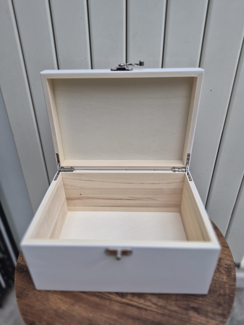 May include: An open, white wooden box with a silver clasp and hinges. The interior of the box is natural wood, contrasting with the painted exterior. The box is sitting on a wooden surface.