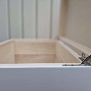 May include: An open, white wooden box with a light-colored interior. The box has a hinged lid, revealing the inside. The box is rectangular and appears to be empty. The hinges are silver-colored.