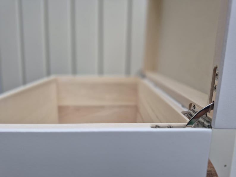 May include: An open, white wooden box with a light-colored interior. The box has a hinged lid, revealing the inside. The box is rectangular and appears to be empty. The hinges are silver-colored.