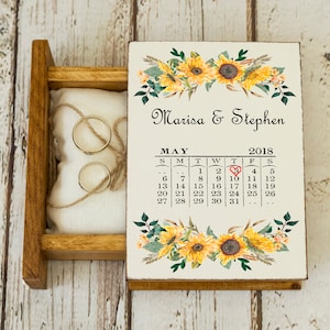 Puede incluir: Una caja de madera para anillos con un diseño de girasol y un calendario en la tapa. El calendario muestra la fecha 10 de mayo de 2018, con un corazón rojo marcando la fecha. La caja está abierta, revelando una almohada blanca con dos anillos de oro.
