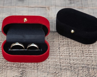 May include: Two gold wedding bands with small blue stones are displayed in a red velvet ring box. A black velvet ring box is also shown in the image.