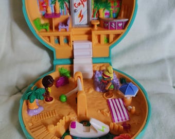 Könnte beinhalten: Ein geöffnetes, orangefarbenes und türkisfarbenes Polly Pocket Spielset im Vintage-Stil. Das Spielset zeigt eine Strandszene mit Miniaturfiguren, Surfbrettern, einem Café und einem Boot. Der Innenraum ist detailliert mit kleinem Zubehör und einem Strandthema.