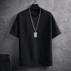 Puede incluir: Una camiseta negra de manga corta con una superficie texturizada cuelga de una percha negra. Un collar de cadena plateada con un colgante rectangular est&aacute; drapeado sobre la camisa. El colgante tiene una serie de peque&ntilde;os dise&ntilde;os cuadrados.