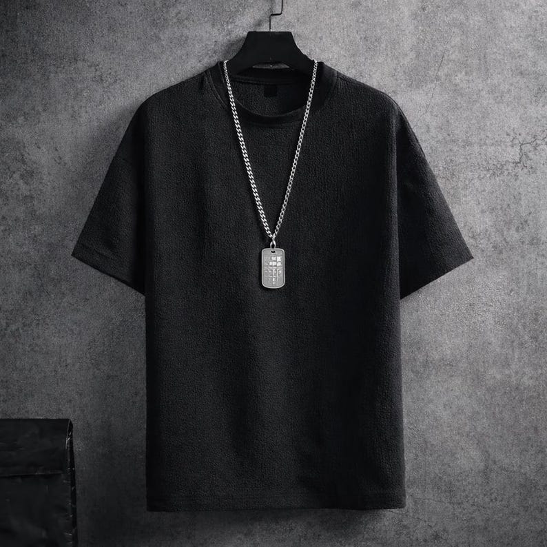 Puede incluir: Una camiseta negra de manga corta con una superficie texturizada cuelga de una percha negra. Un collar de cadena plateada con un colgante rectangular est&aacute; drapeado sobre la camisa. El colgante tiene una serie de peque&ntilde;os dise&ntilde;os cuadrados.