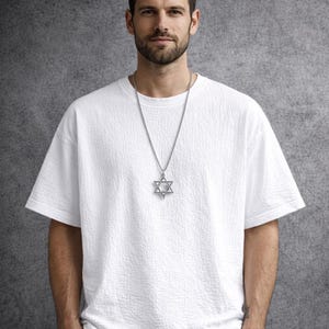 Puede incluir: Un collar con colgante de plata con una estrella de David. El colgante es una estrella de seis puntas, suspendida de una delicada cadena. El collar se lleva sobre una camiseta blanca texturizada.