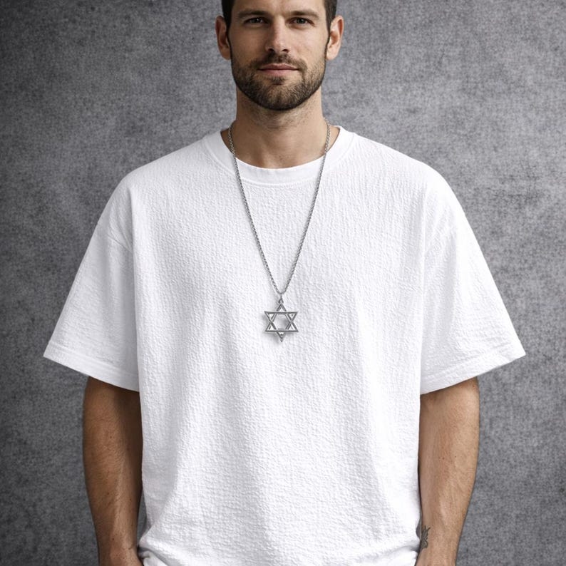 Puede incluir: Un collar con colgante de plata con una estrella de David. El colgante es una estrella de seis puntas, suspendida de una delicada cadena. El collar se lleva sobre una camiseta blanca texturizada.