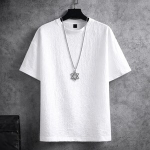 Puede incluir: Una camiseta blanca texturizada con mangas cortas cuelga de una percha negra. Un collar plateado con un colgante de estrella de David est&aacute; drapeado sobre la camisa. El fondo es una pared gris.