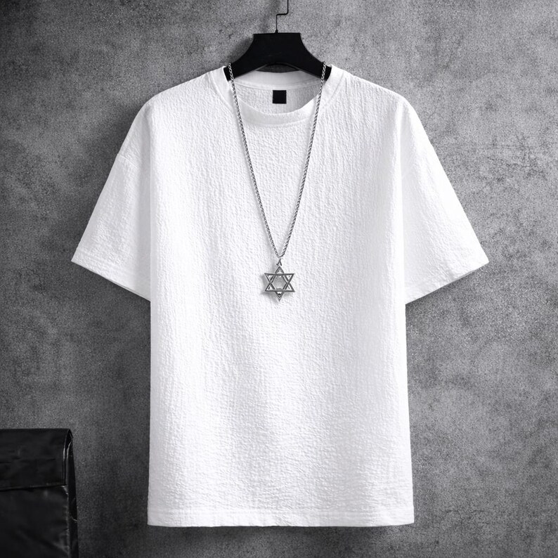 Puede incluir: Una camiseta blanca texturizada con mangas cortas cuelga de una percha negra. Un collar plateado con un colgante de estrella de David est&aacute; drapeado sobre la camisa. El fondo es una pared gris.