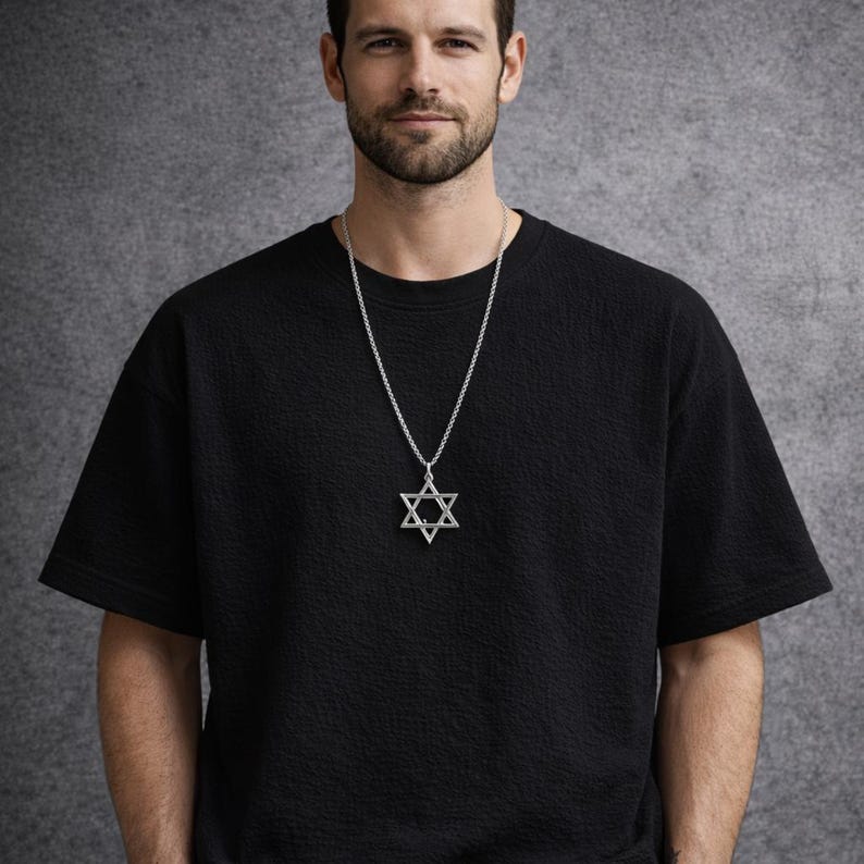 Puede incluir: Un collar con un colgante de estrella de David plateado. El colgante cuelga de una fina cadena. El collar se lleva sobre una camiseta negra texturizada. El fondo es gris liso.