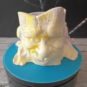 Op de afbeelding: Een crèmekleurige keramische plantenbak met Frankenstein-thema, met gele en blauwe spikkels. De plantenbak heeft de vorm van het hoofd van Frankenstein, met een platte bovenkant en een ronde basis. De plantenbak staat op een blauw oppervlak.
