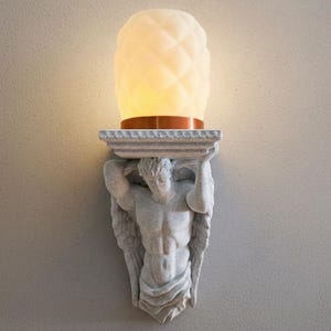 May include: A wall-mounted light fixture with a classical male figure supporting a shelf. The shelf holds a glowing, textured, white glass light with a copper-coloured base. The sconce is mounted on a neutral-coloured wall.