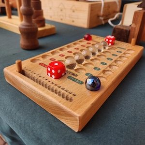 May include: A wooden board game with a series of holes and a scoring track. The game includes red dice and clear glass marbles. The board is made of light-colored wood and has the words "SCORE" and "START" printed on it.