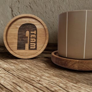 Könnte beinhalten: Ein runder Holzuntersetzer mit dem eingravierten Wort "TEAM". Ein brauner Becher mit weißen vertikalen Streifen steht auf einem passenden Untersetzer. Die Gegenstände befinden sich auf einer Holzoberfläche.