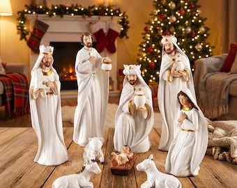 May include: A Christmas nativity scene featuring figurines of the three wise men, Mary, Joseph, baby Jesus, and animals. The figures are white with gold accents, set on a wooden surface. A Christmas tree and fireplace are in the background.
