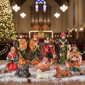 May include: A Christmas nativity scene with figures in colorful robes. The set includes Mary, Joseph, the three wise men, baby Jesus in a manger, and animals. The scene is set on a table with a Christmas tree in the background.