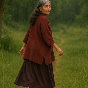 May include: A woman wearing a burgundy-colored top and a dark purple skirt walks on a path through a green field. The top has three-quarter sleeves and a split hem. She wears brown shoes.