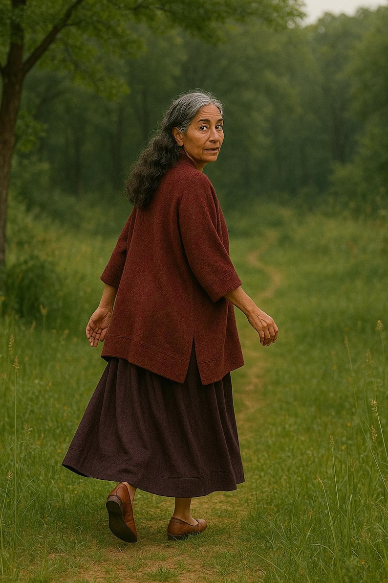 May include: A woman wearing a burgundy-colored top and a dark purple skirt walks on a path through a green field. The top has three-quarter sleeves and a split hem. She wears brown shoes.