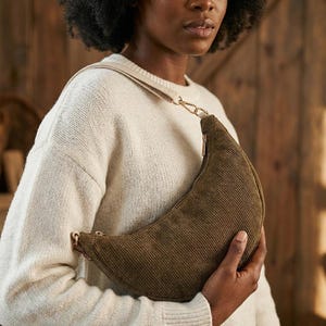 May include: A crescent-shaped, olive green corduroy shoulder bag with a gold chain strap. The bag has a zipper closure and a beige strap. The bag is held by a person wearing a cream-colored sweater.
