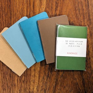 Può includere: Una collezione di piccoli quaderni in vari colori, tra cui beige, azzurro, turchese, marrone e verde. Il quaderno verde è confezionato e ha un'etichetta bianca con la scritta "THE MICRO NOTEBOOK 16 PAGES - PLAIN 6.35 x 8.89 cm HANDMADE."