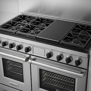 May include: Stainless steel Wolf range with six burners, a griddle, and two ovens. The range features black burner grates and control knobs. The Wolf logo is visible on the oven doors. A modern kitchen appliance.