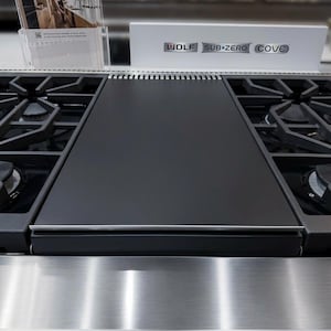 May include: A close-up of a stainless steel gas range with black burners and a black cover. The brand name "WOLF SUB-ZERO COVE" is displayed on a white sign above the range. A brochure is visible on the left.