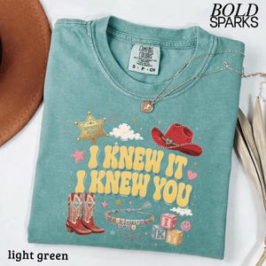 Może przedstawiać: Jasnozielony t-shirt z nadrukiem graficznym z ż&oacute;łtym napisem "I KNEW IT I KNEW YOU". Wz&oacute;r zawiera złotą gwiazdę szeryfa, czerwony kowbojski kapelusz, dwa kowbojskie buty, bransoletkę z koralik&oacute;w oraz klocki z literami. Na koszulce leży srebrny naszyjnik.